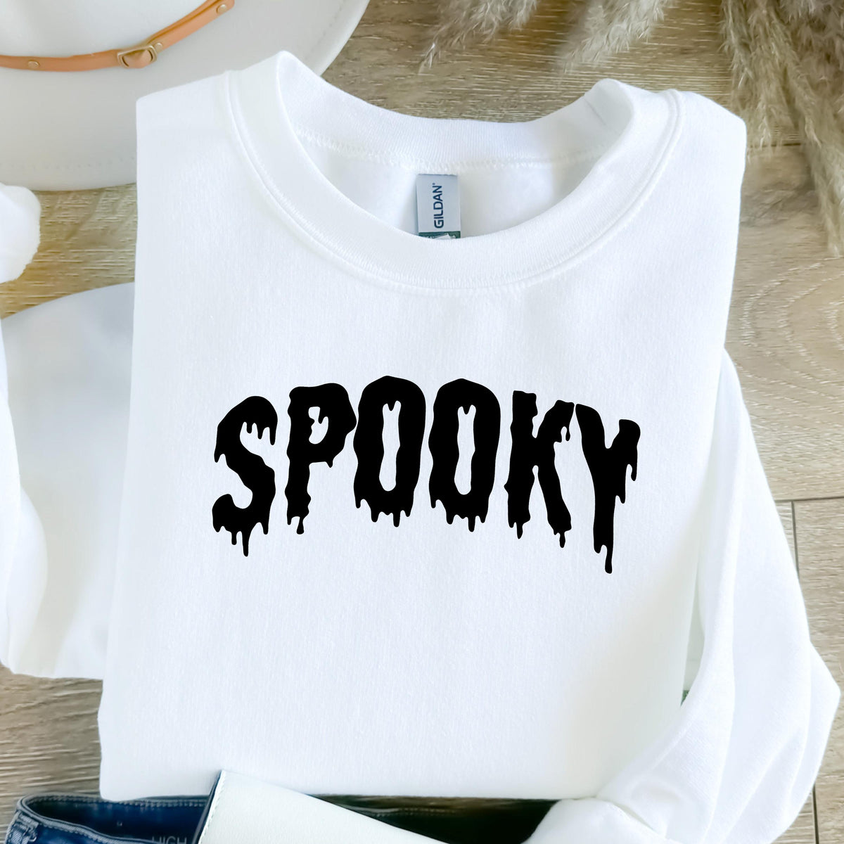 May include: A white long-sleeve crewneck sweatshirt featuring the word SPOOKY printed in a black, dripping, liquid-style font across the chest. The garment has a standard ribbed neckline and a Gildan brand label visible inside the collar.