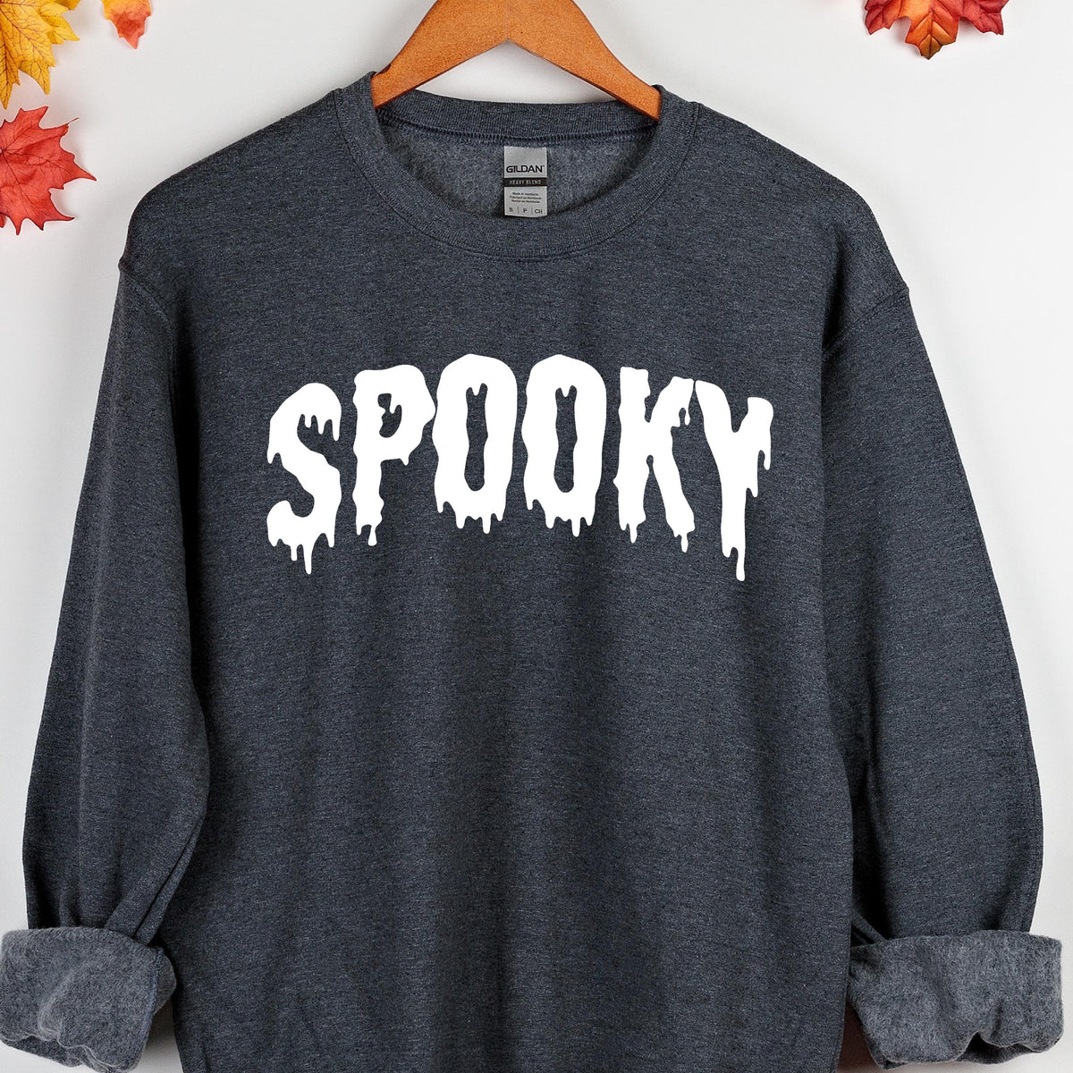 May include: A dark heather gray crewneck sweatshirt featuring the word SPOOKY printed in large white letters with a dripping, melted effect. The garment has long sleeves with ribbed cuffs and a ribbed neckline, displayed on a wooden hanger.