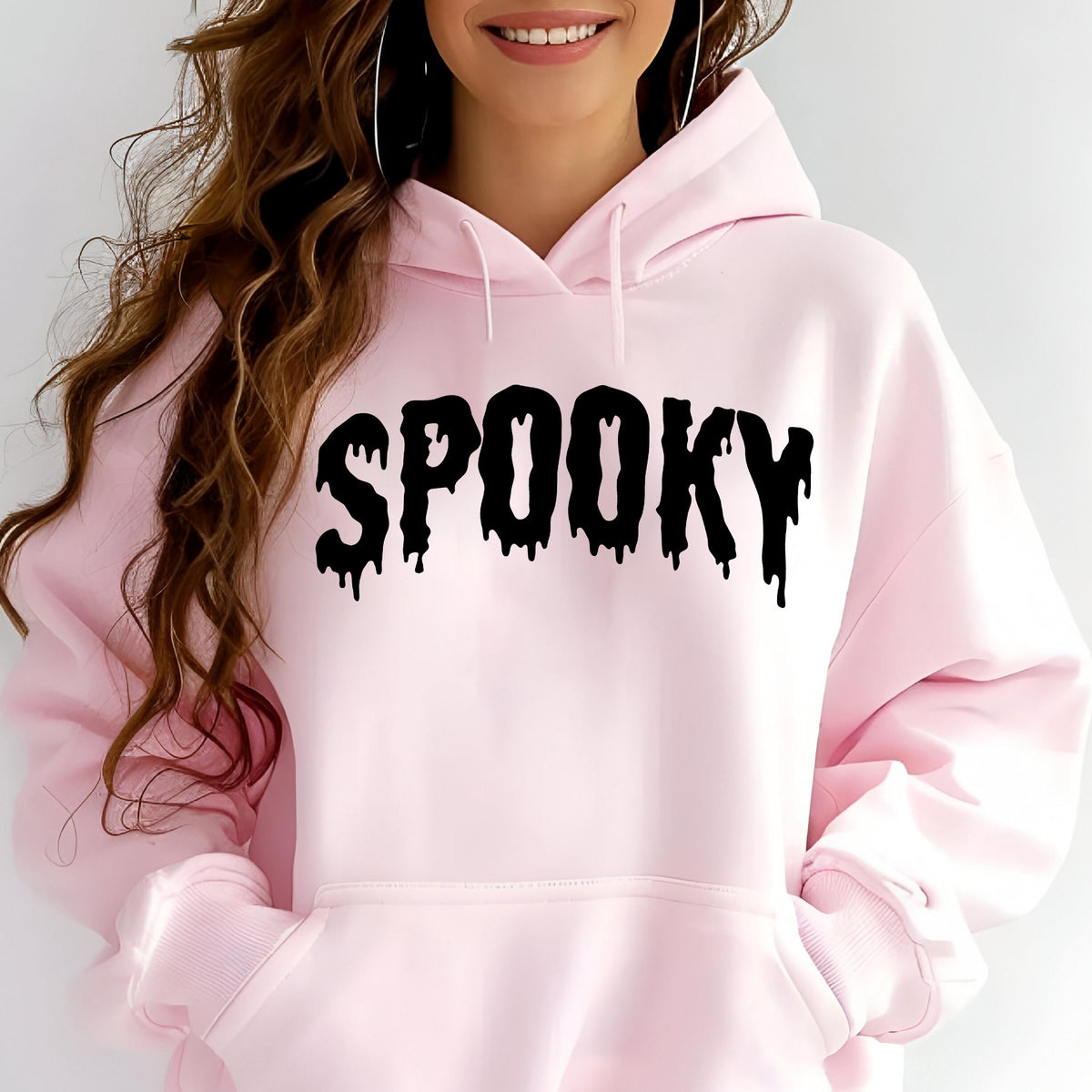 May include: A light pink hooded sweatshirt featuring a front kangaroo pocket and long sleeves. The word SPOOKY is printed across the chest in a black, dripping, stylized font. The garment has a hood with matching pink drawstrings.