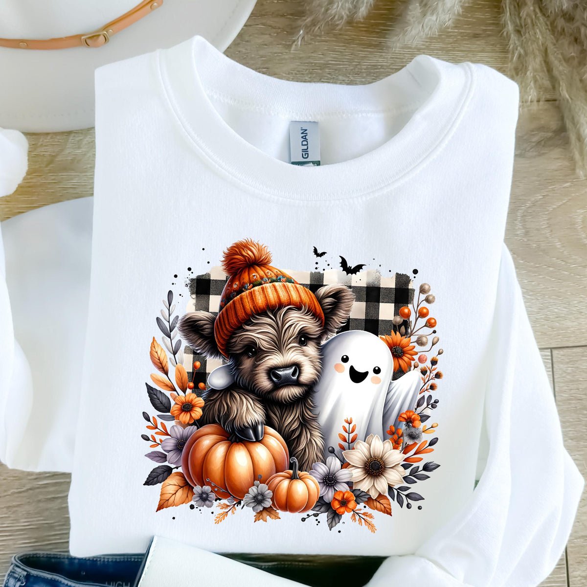 May include: A white crewneck sweatshirt featuring a graphic print of a fluffy brown highland cow wearing an orange knit beanie, standing next to a white ghost, a large orange pumpkin, and autumn flowers against a black and white buffalo plaid.