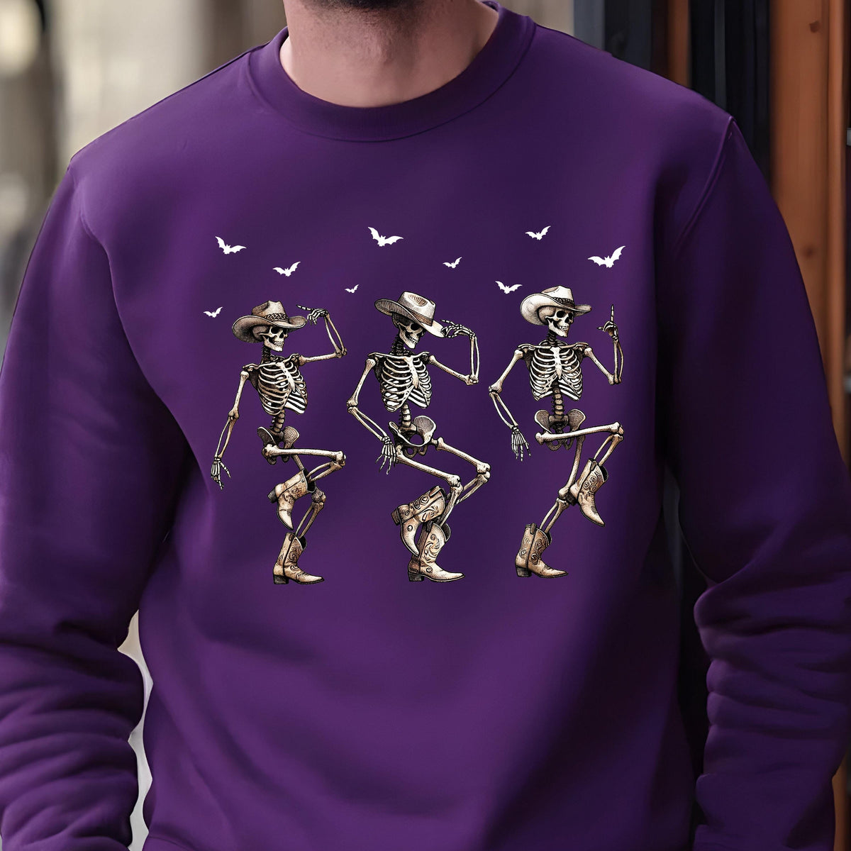 May include: A purple sweatshirt featuring a graphic of three dancing skeletons wearing cowboy hats and boots. White bats are scattered across the top. The skeletons are in various dance poses.