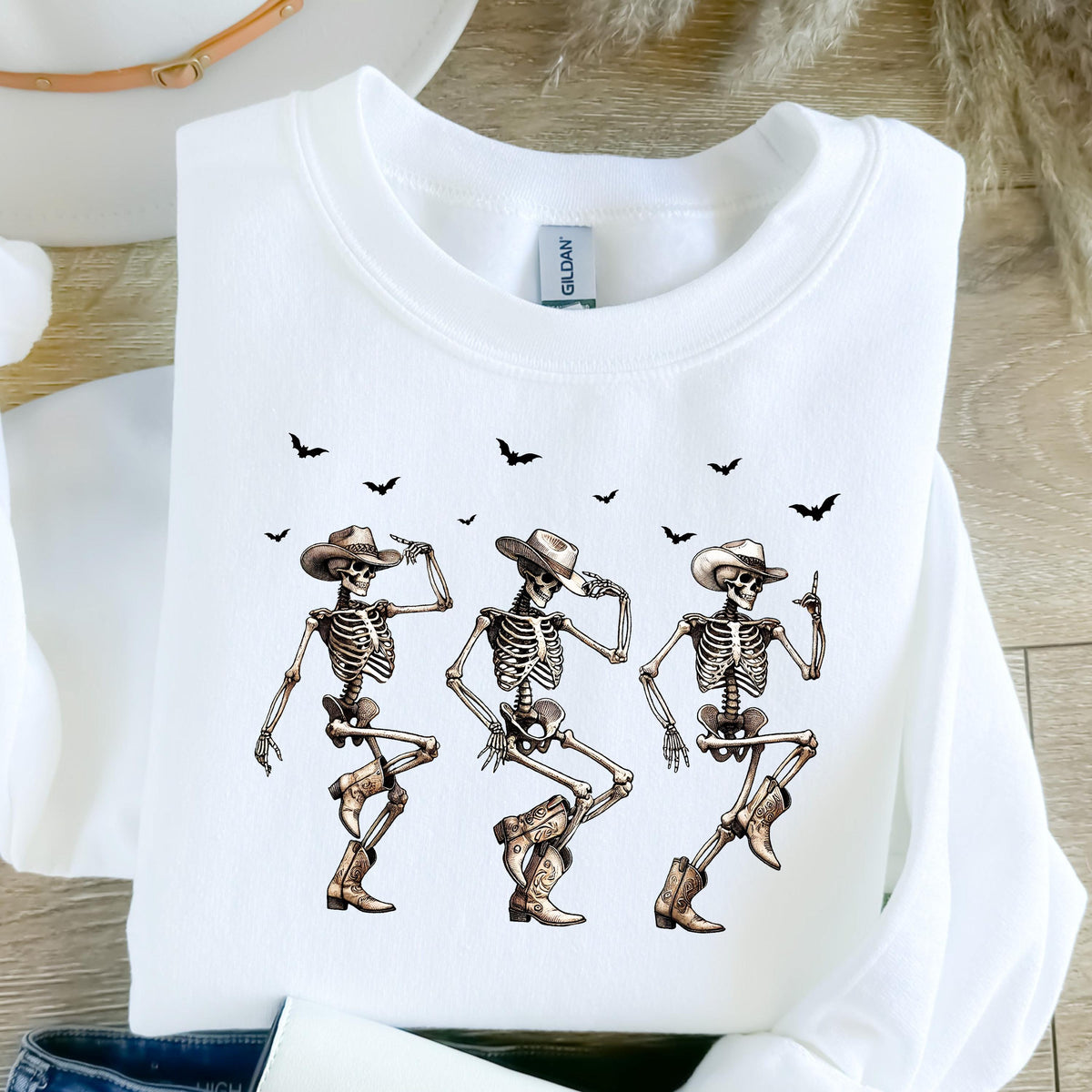 May include: White sweatshirt featuring a graphic of three dancing skeletons wearing cowboy hats and boots. Black bat silhouettes are scattered above the skeletons. The design has a Western and Halloween theme.