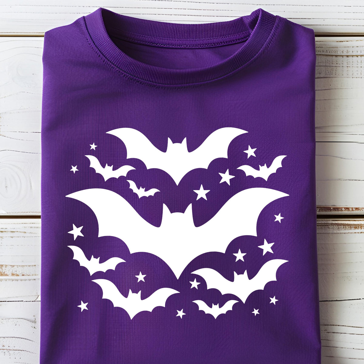 May include: A folded purple t-shirt featuring a white bat and star design. The graphic includes several bats of varying sizes and multiple five-pointed stars. The shirt is made of a soft material and is suitable for Halloween.