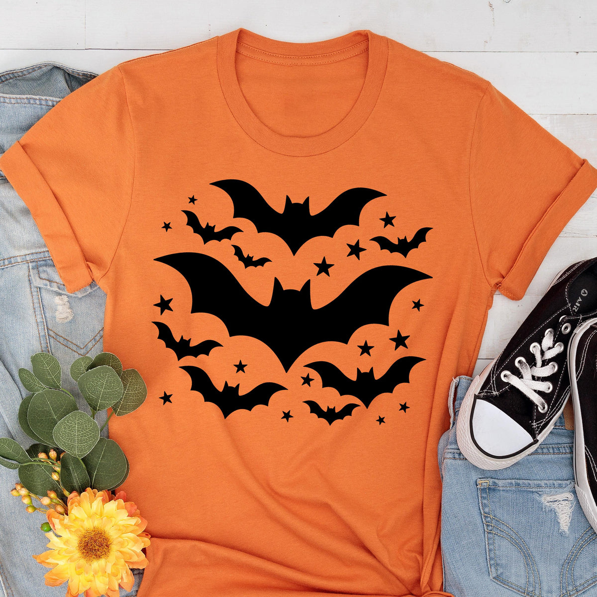 May include: Orange t-shirt featuring a black bat design with stars. The graphic includes several bats of varying sizes and stars, creating a Halloween theme. The shirt is paired with denim and black sneakers.