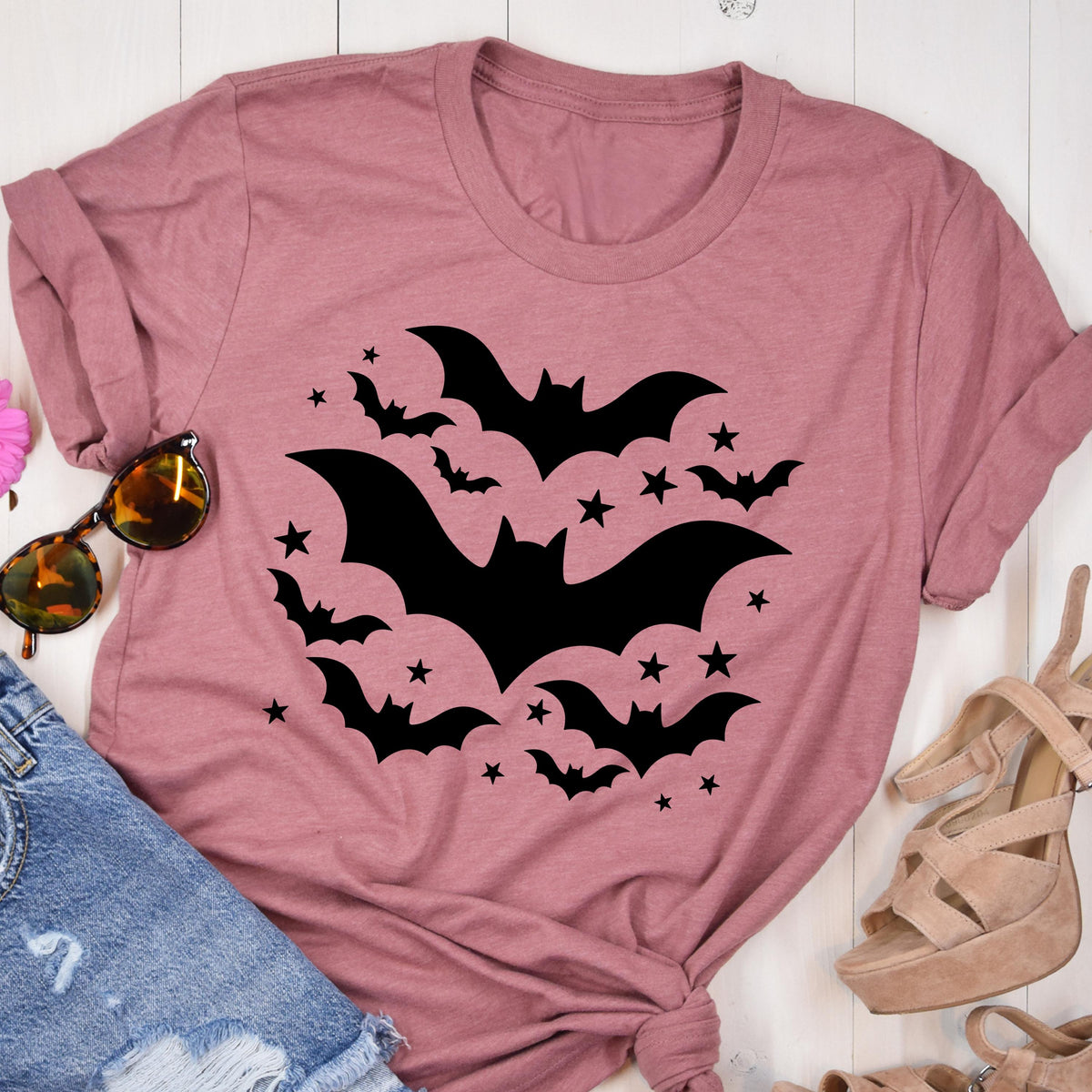 May include: Dusty rose t-shirt featuring a black bat and star design. The graphic includes several bats of varying sizes and stars arranged in a circular pattern. The shirt is folded, with sunglasses and denim shorts visible.