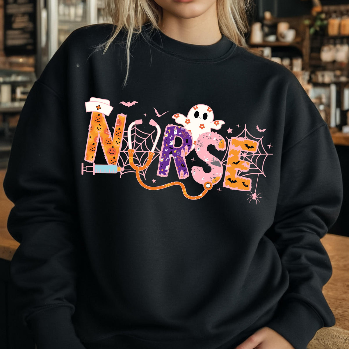 May include: Black sweatshirt with a colorful Halloween-themed "NURSE" graphic. The design includes a syringe, stethoscope, spiderwebs, bats, and a ghost. The letters are orange, purple, and pink.