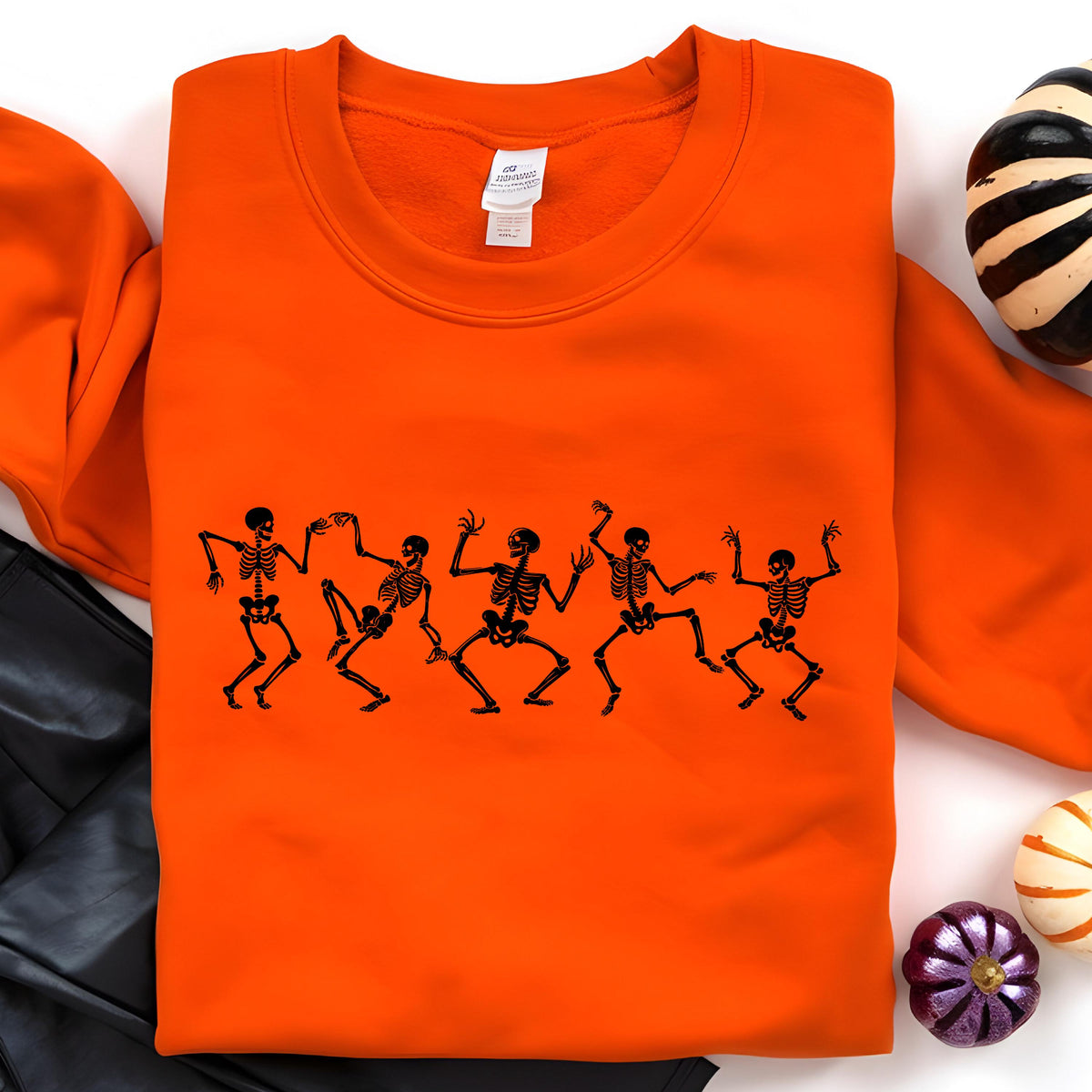 May include: Bright orange sweatshirt featuring a black graphic of dancing skeletons. The crew neck sweatshirt is a Halloween-themed apparel item. The design shows five skeletons in various dance poses, perfect for spooky season.