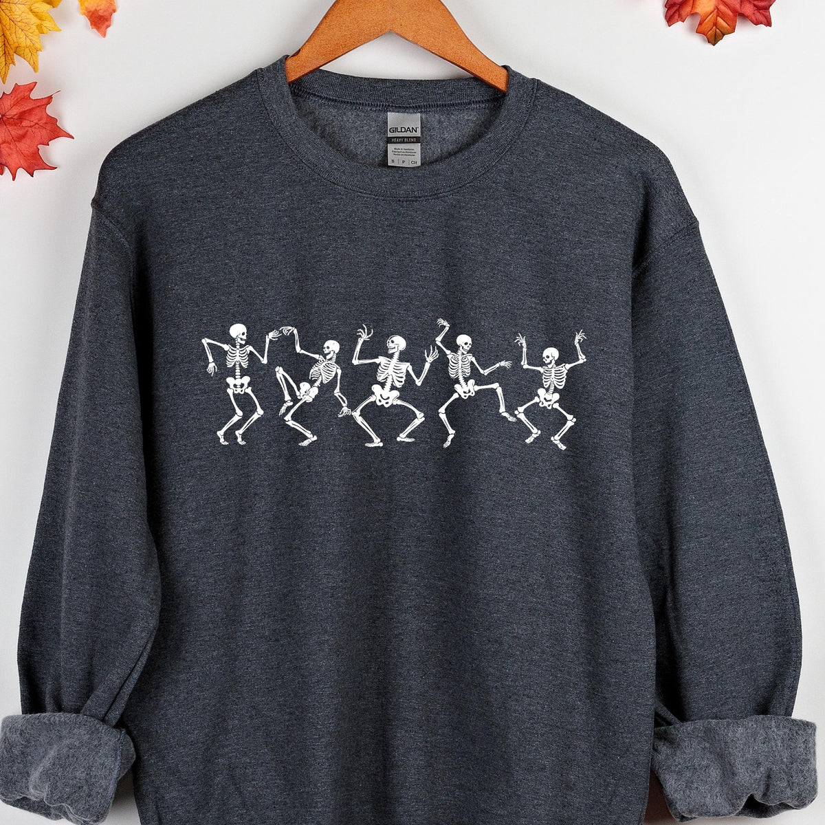 May include: Dark gray sweatshirt featuring a white graphic of dancing skeletons. The skeletons are in a line, with their arms and legs in various positions, giving the impression of movement. The sweatshirt has a crew neck and long sleeves, perfect for Halloween.