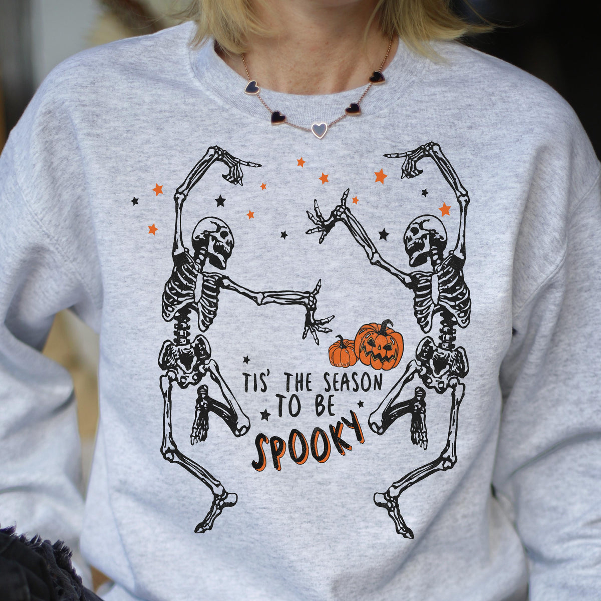 May include: A light gray sweatshirt featuring a Halloween design. The graphic includes two dancing skeletons, orange pumpkins, and stars. The text reads "Tis' the season to be spooky" in black and orange.