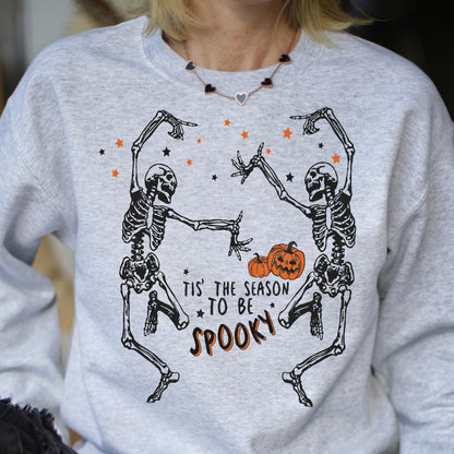 May include: A light gray sweatshirt featuring a Halloween design. The graphic includes two dancing skeletons, orange pumpkins, and stars. The text reads "Tis' the season to be spooky" in black and orange.