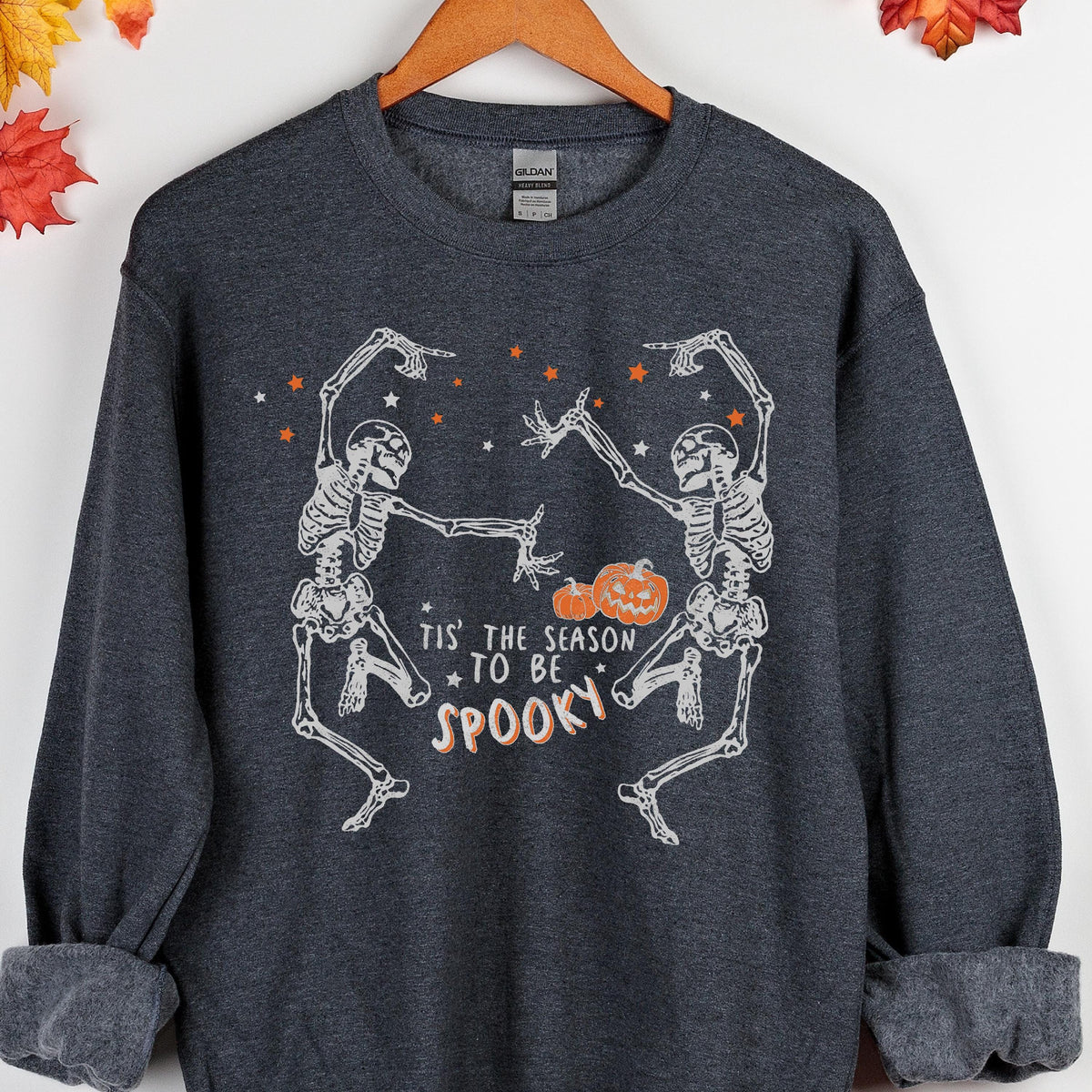 May include: Dark gray sweatshirt with a Halloween-themed graphic. The design features two dancing skeletons, orange stars, and pumpkins. The text reads "Tis the season to be spooky" in a playful font. The sweatshirt has a crew neck and long sleeves.