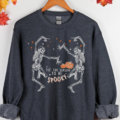 May include: Dark gray sweatshirt with a Halloween-themed graphic. The design features two dancing skeletons, orange stars, and pumpkins. The text reads "Tis the season to be spooky" in a playful font. The sweatshirt has a crew neck and long sleeves.
