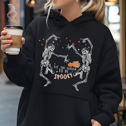 May include: Black hoodie featuring a Halloween design. The graphic includes two dancing skeletons, a jack-o'-lantern, and the text "Tis' the Season to be Spooky" in white and orange. The hoodie has a drawstring hood and a kangaroo pocket.