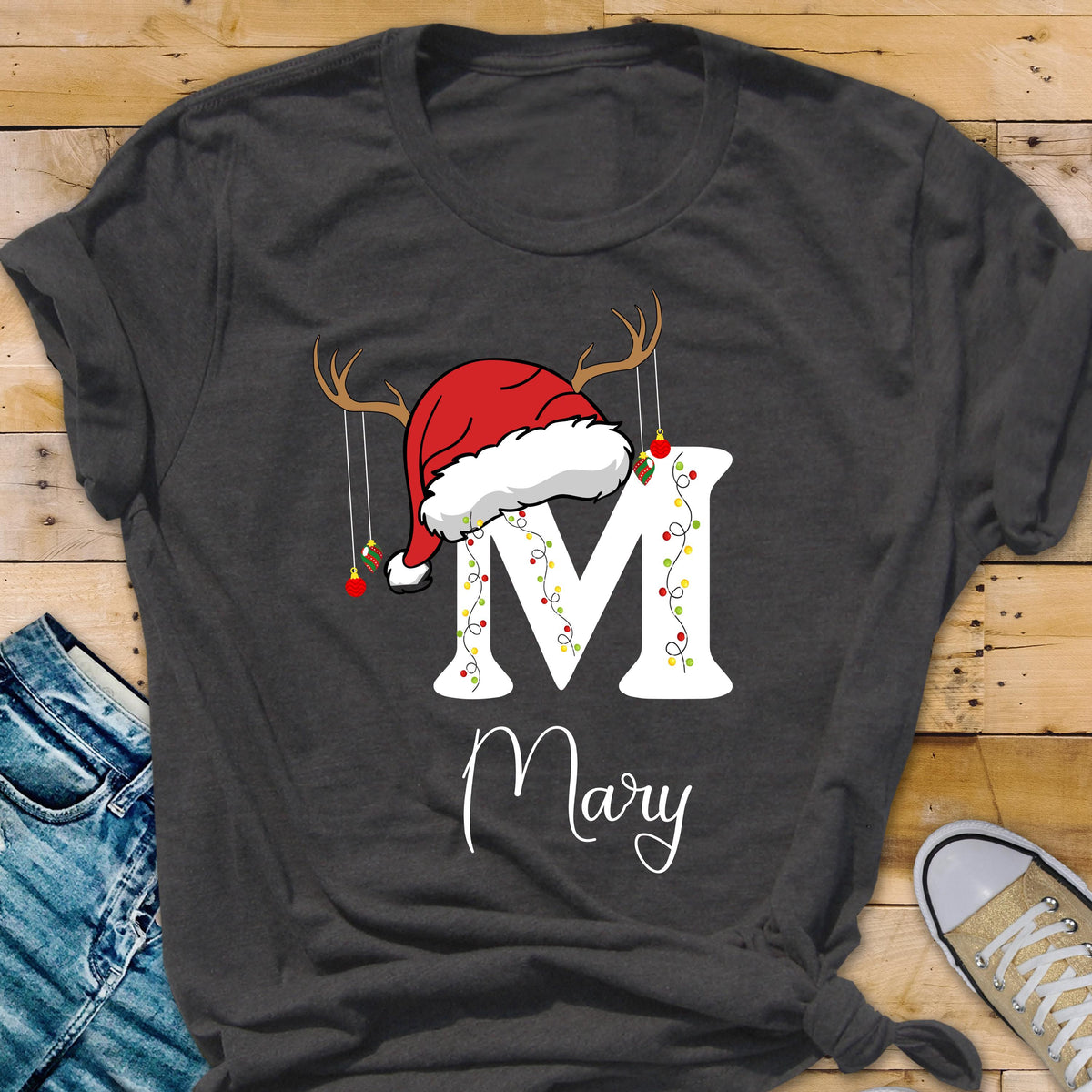 May include: A dark gray short-sleeve t-shirt featuring a large white letter M decorated with colorful Christmas lights. A red Santa hat with brown reindeer antlers sits atop the letter, with small hanging ornaments. The name Mary is printed below.