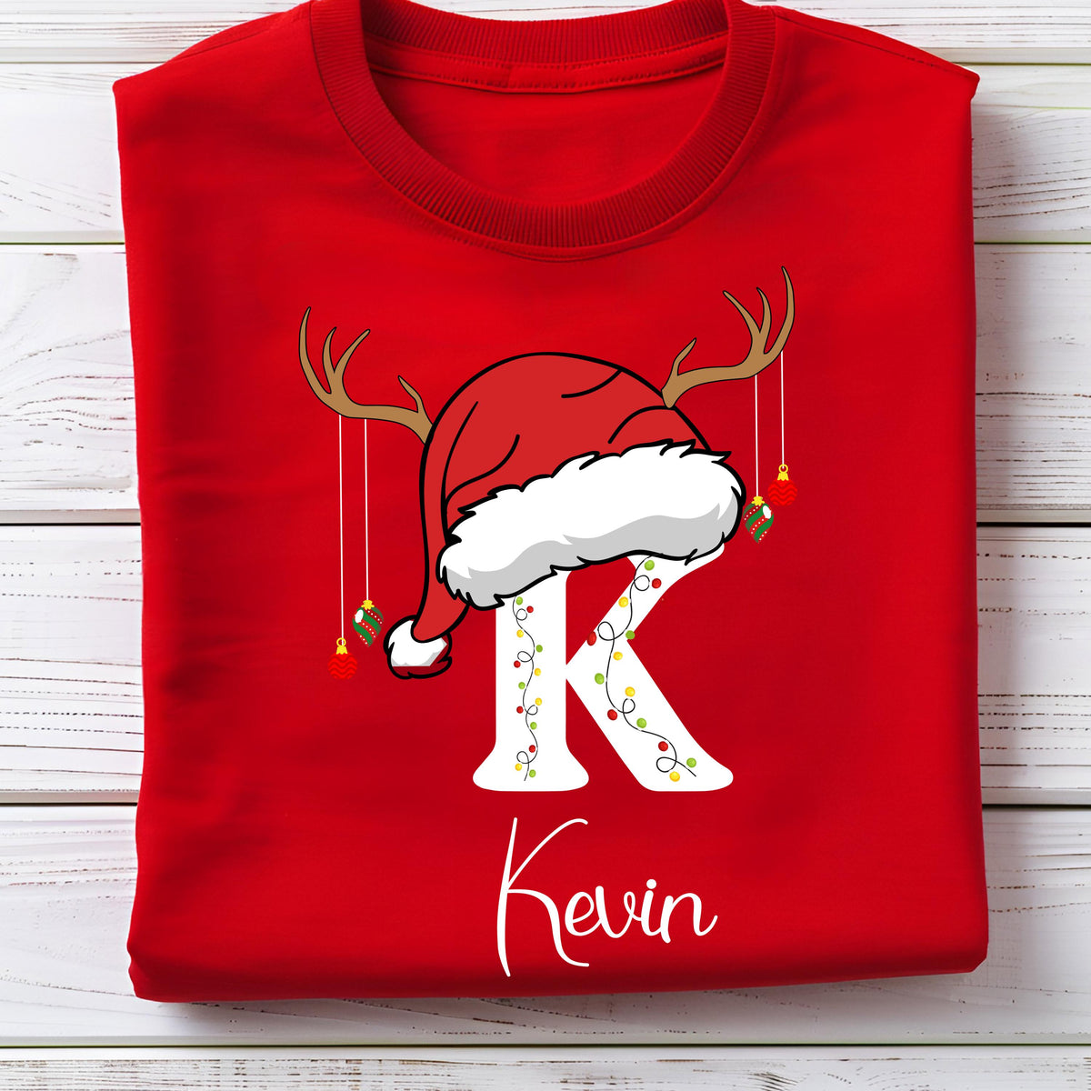 May include: A bright red t-shirt featuring a large white letter K decorated with colorful string lights. The letter is topped with a red Santa hat and brown reindeer antlers, with small hanging Christmas ornaments and the name Kevin printed below.