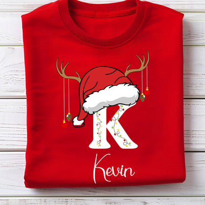 May include: A bright red t-shirt featuring a large white letter K decorated with colorful string lights. The letter is topped with a red Santa hat and brown reindeer antlers, with small hanging Christmas ornaments and the name Kevin printed below.