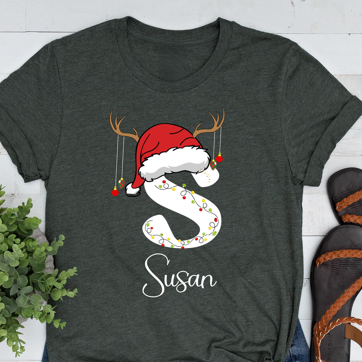 May include: A dark charcoal gray t-shirt featuring a large white letter S decorated with colorful Christmas lights. The letter is topped with a red Santa hat and brown reindeer antlers with hanging red ornaments. The name Susan is printed below.