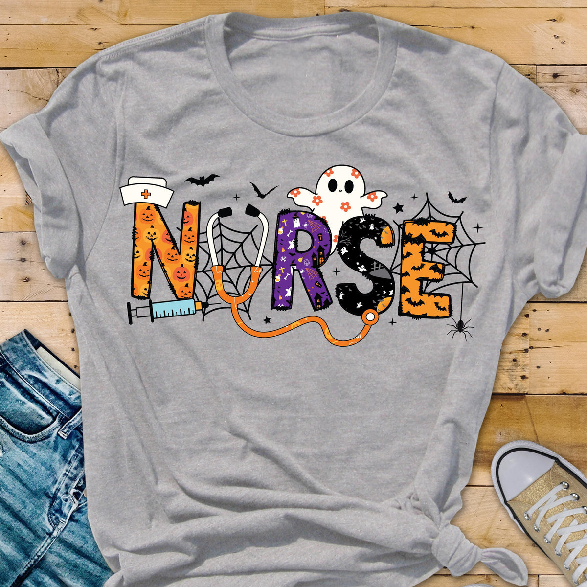 May include: A gray t-shirt featuring the word "NURSE" in a Halloween-themed design. The letters are decorated with pumpkins, ghosts, and spiderwebs. A stethoscope and syringe are also included in the design.