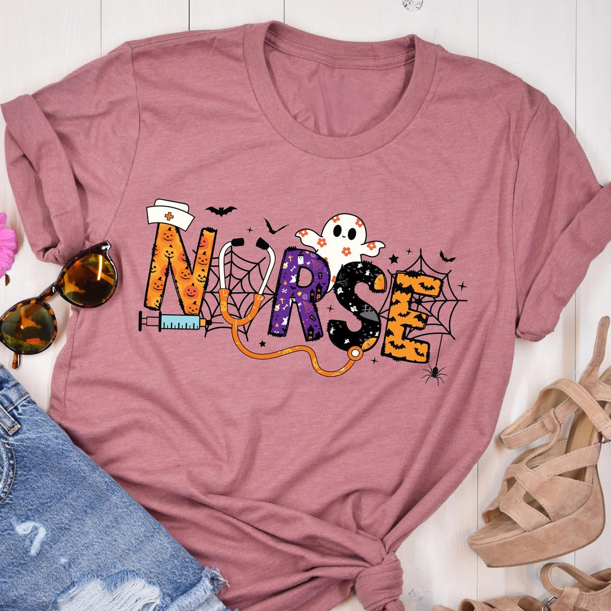May include: Dusty rose t-shirt with the word "NURSE" in a Halloween-themed design. The letters are decorated with pumpkins, spiderwebs, and a ghost. A stethoscope and syringe are also included in the design.