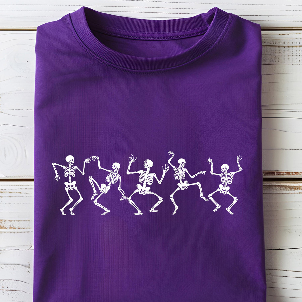 May include: A purple t-shirt featuring a white graphic of six dancing skeletons. The skeletons are in various poses, with their arms and legs outstretched. The shirt is folded neatly on a white wooden surface.