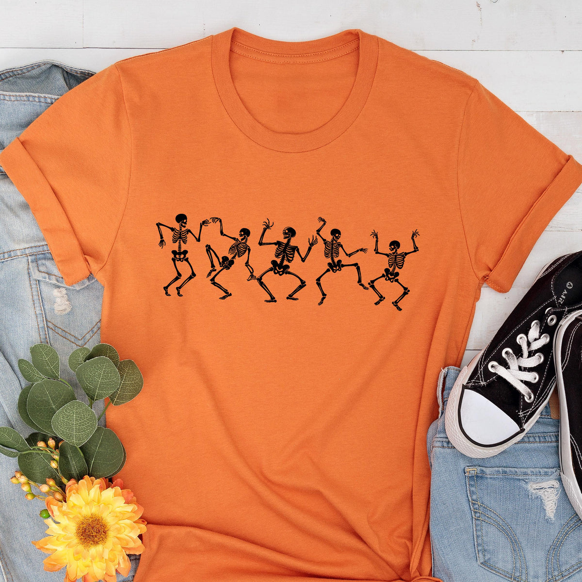 May include: An orange t-shirt featuring a black graphic of dancing skeletons. The skeletons are in various poses, with arms and legs outstretched. The shirt is made of a soft material and has a classic crew neck design.