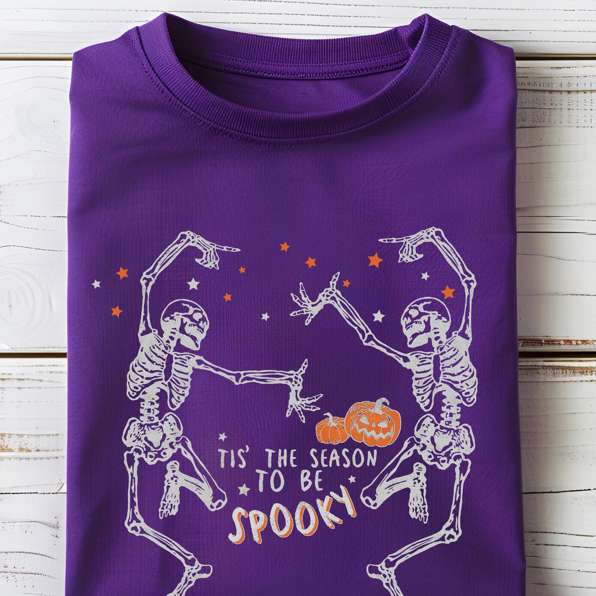 May include: A purple t-shirt featuring a Halloween design. The graphic includes two dancing skeletons, orange stars, and pumpkins. The text reads "Tis' the season to be spooky."