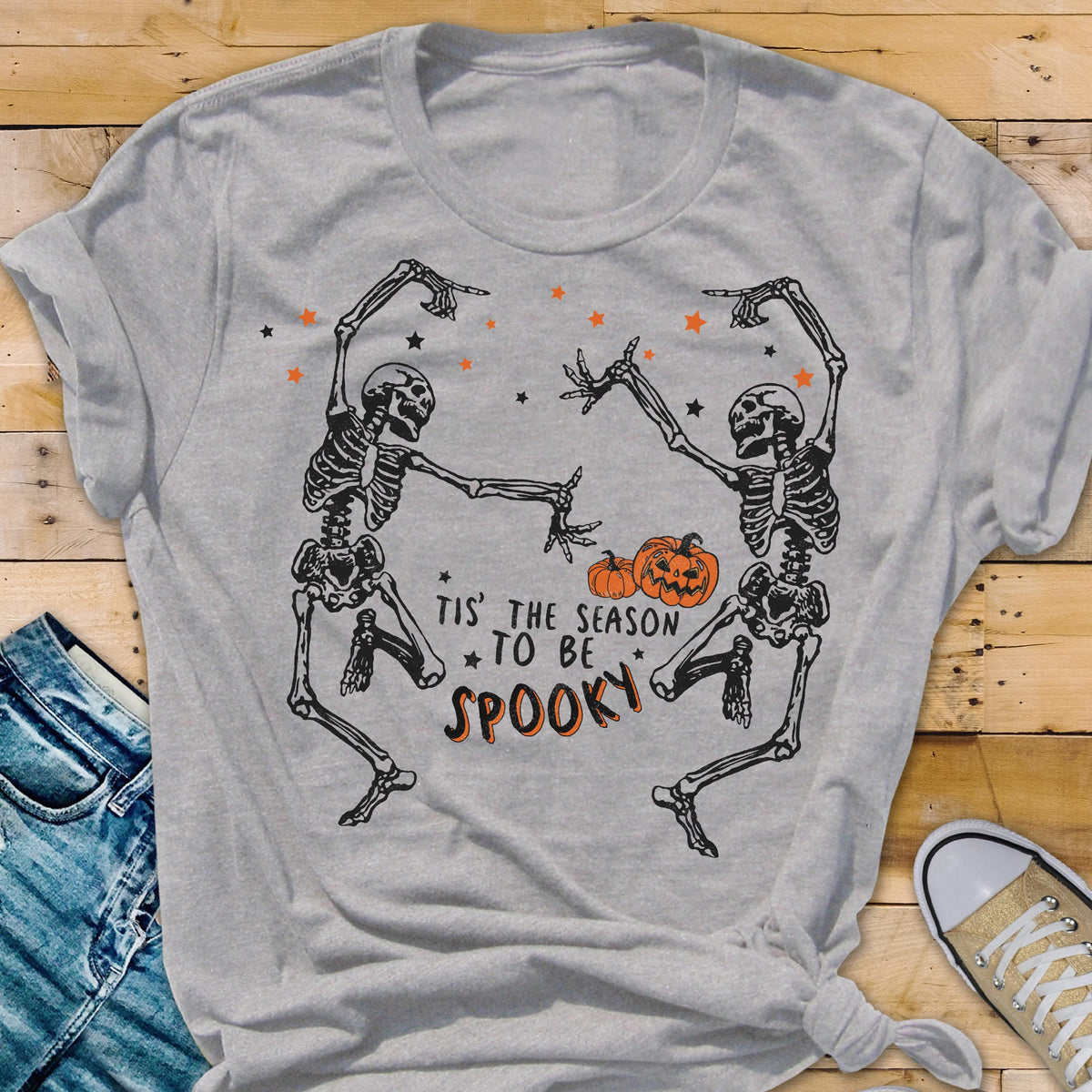 May include: A light gray t-shirt featuring a Halloween design. Two dancing skeletons are depicted, with orange stars and pumpkins. The text reads "Tis' the season to be spooky" in orange and black.