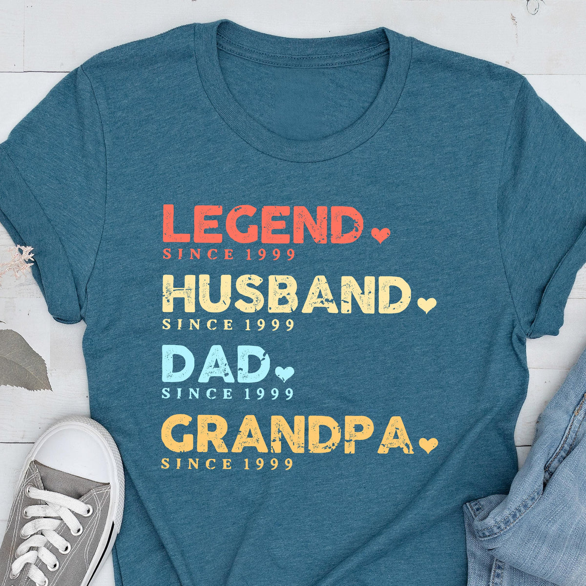 May include: Teal t-shirt with the words "LEGEND, HUSBAND, DAD, GRANDPA" in red, yellow, and blue text. Each word is followed by "SINCE 1999". A gray sneaker is visible in the lower left corner.