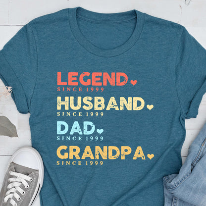 May include: Teal t-shirt with the words "LEGEND, HUSBAND, DAD, GRANDPA" in red, yellow, and blue text. Each word is followed by "SINCE 1999". A gray sneaker is visible in the lower left corner.