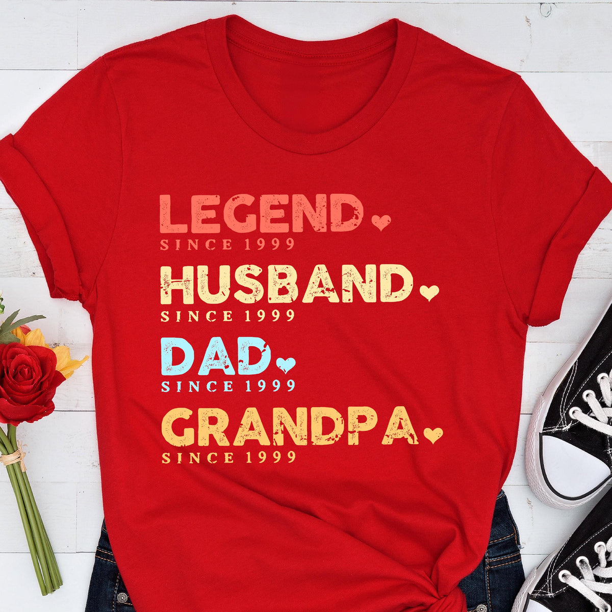 May include: A red t-shirt with the words "LEGEND, HUSBAND, DAD, GRANDPA" in various colors, each followed by "SINCE 1999". The shirt has rolled-up sleeves and is paired with blue jeans and black sneakers.