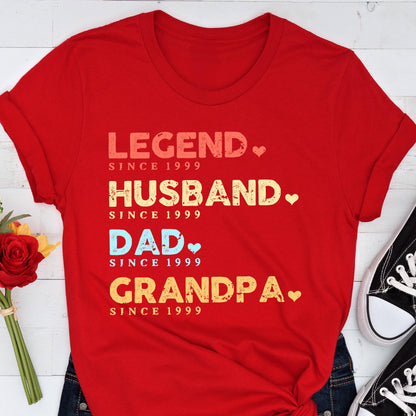 May include: A red t-shirt with the words "LEGEND, HUSBAND, DAD, GRANDPA" in various colors, each followed by "SINCE 1999". The shirt has rolled-up sleeves and is paired with blue jeans and black sneakers.