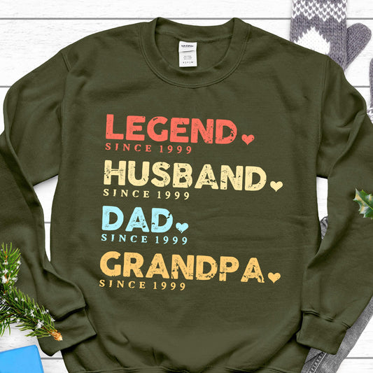 May include: An olive green sweatshirt with the words "LEGEND," "HUSBAND," "DAD," and "GRANDPA" in various colors. Each word is followed by "SINCE 1999" and a heart symbol. A casual, comfortable garment.