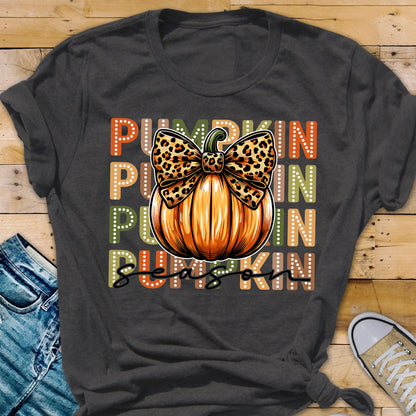 May include: Dark gray t-shirt featuring a large orange pumpkin with a leopard print bow. The word "PUMPKIN" is stacked vertically in orange, green, and brown. The words "PUMPKIN SEASON" are written in a script font.