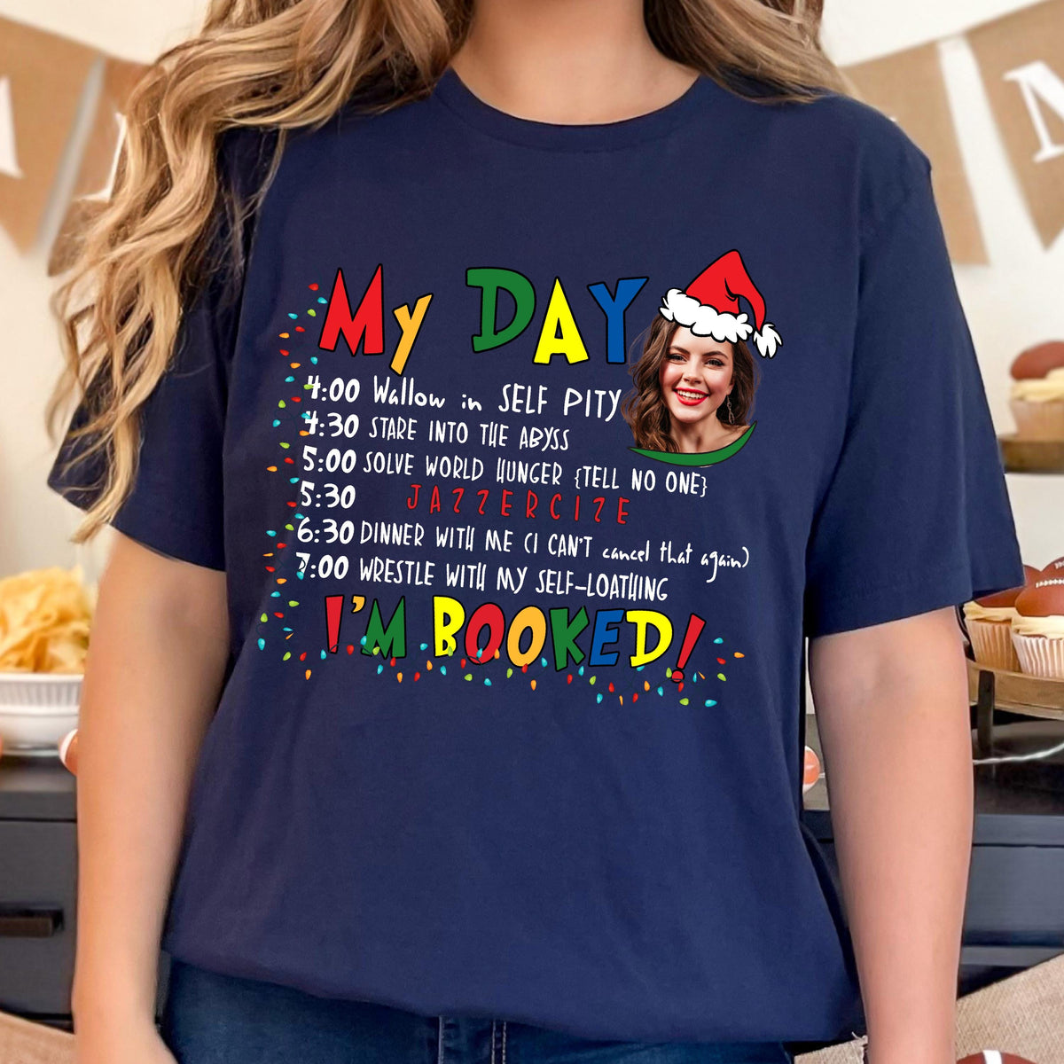 May include: Navy blue t-shirt with the text "My Day" in colorful letters. The shirt features a list of humorous activities, including "Wallow in Self Pity" and "Jazzercise." A photo of a woman wearing a Santa hat is also on the shirt. The words "I'm Booked!" are at the bottom.
