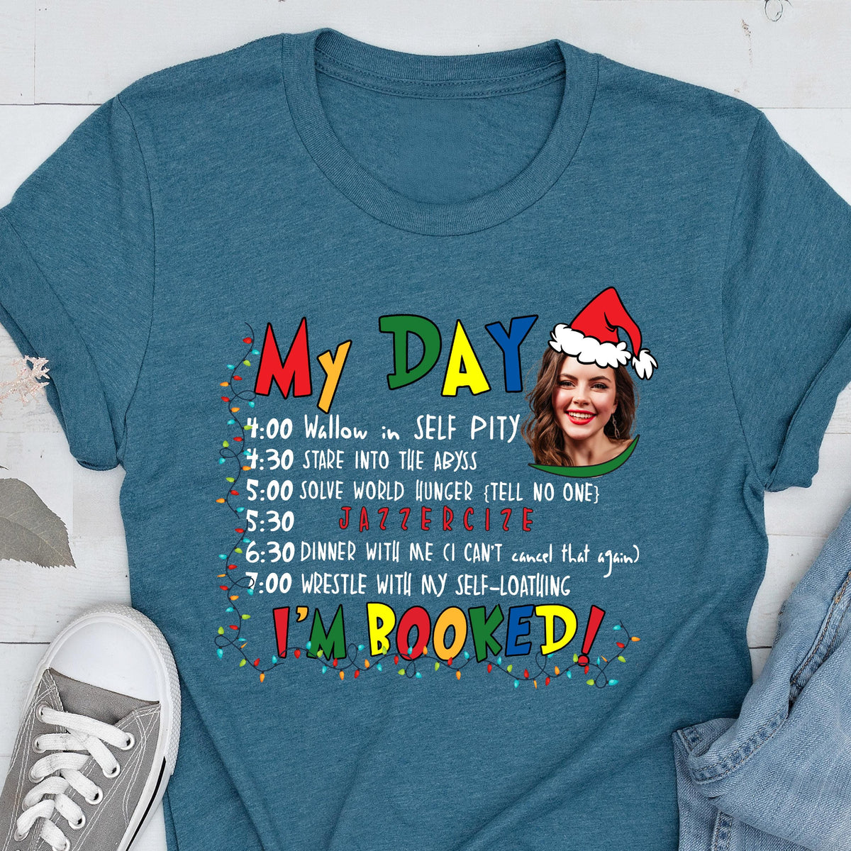 May include: Teal t-shirt with the text "My Day" and a schedule of humorous activities. The schedule includes "Wallow in SELF PITY," "Stare into the Abyss," and "JAZZERCIZE." A woman wearing a Santa hat is in the image.