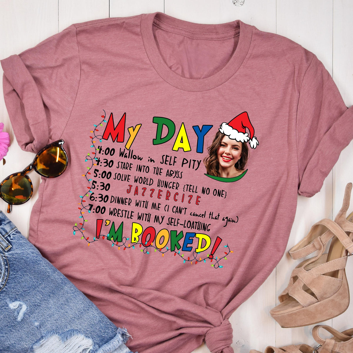 May include: A mauve t-shirt with the words "My Day" in colorful letters. The shirt features a humorous schedule including "Wallow in Self Pity" and "Jazzerize." A woman wearing a Santa hat is also pictured.