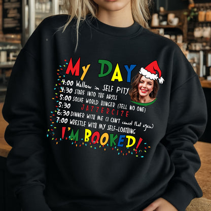 May include: A black crewneck sweatshirt featuring a colorful graphic with the text My Day and I'm Booked, a list of activities including Wallow in Self Pity, Stare into the Abyss, Solve World Hunger, Jazzercize, and a photo of a person wearing a Santa hat.