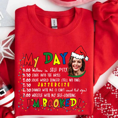 May include: A red crewneck sweatshirt featuring a graphic print with the text My Day followed by a schedule of activities and a photographic portrait of a person wearing a Santa hat. The design includes colorful string lights and text.