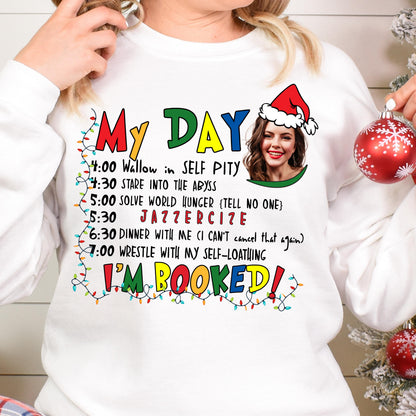May include: A white crewneck sweatshirt featuring a colorful graphic with the text MY DAY and I'M BOOKED, a list of activities, and a custom photo of a person wearing a Santa hat. The design is framed by a string of multi-colored Christmas lights.