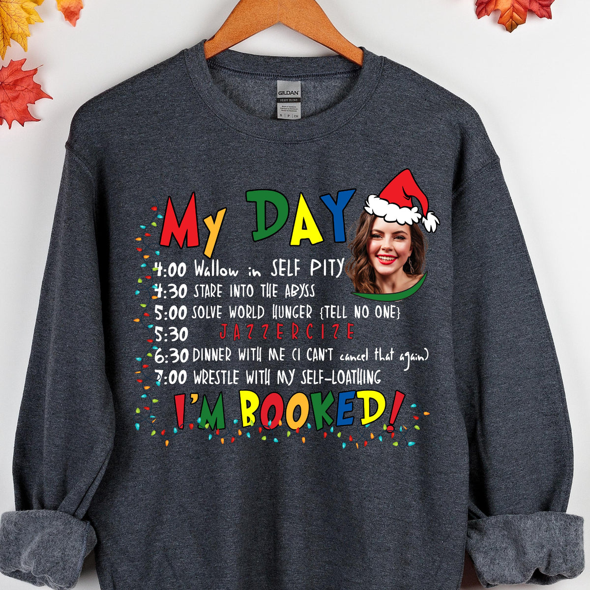 May include: A dark gray long-sleeve sweatshirt featuring a graphic print with the text MY DAY and I'M BOOKED in colorful letters. It includes a schedule of activities and a photographic portrait of a person wearing a red and white Santa hat.