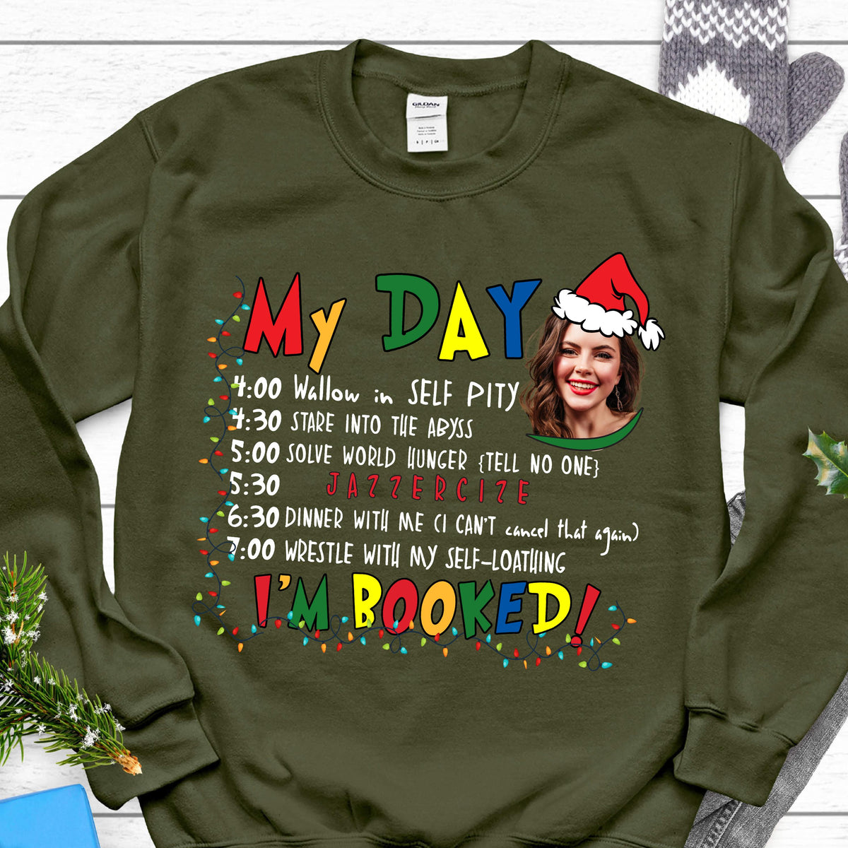 May include: An olive green crewneck sweatshirt featuring a graphic print with the text "MY DAY" and "I'M BOOKED" in colorful letters. The design includes a schedule of activities, a photo of a person wearing a Santa hat, and Christmas lights.