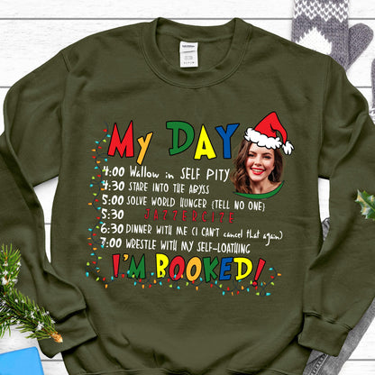 May include: An olive green crewneck sweatshirt featuring a graphic print with the text "MY DAY" and "I'M BOOKED" in colorful letters. The design includes a schedule of activities, a photo of a person wearing a Santa hat, and Christmas lights.