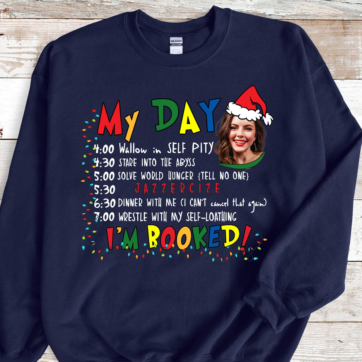 May include: A navy blue crewneck sweatshirt featuring a colorful graphic with the text My Day and I'm Booked, a schedule of activities, and a photographic portrait of a person wearing a red and white Santa hat, surrounded by festive light bulbs.