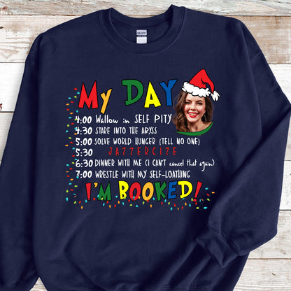 May include: A navy blue crewneck sweatshirt featuring a colorful graphic with the text My Day and I'm Booked, a schedule of activities, and a photographic portrait of a person wearing a red and white Santa hat, surrounded by festive light bulbs.