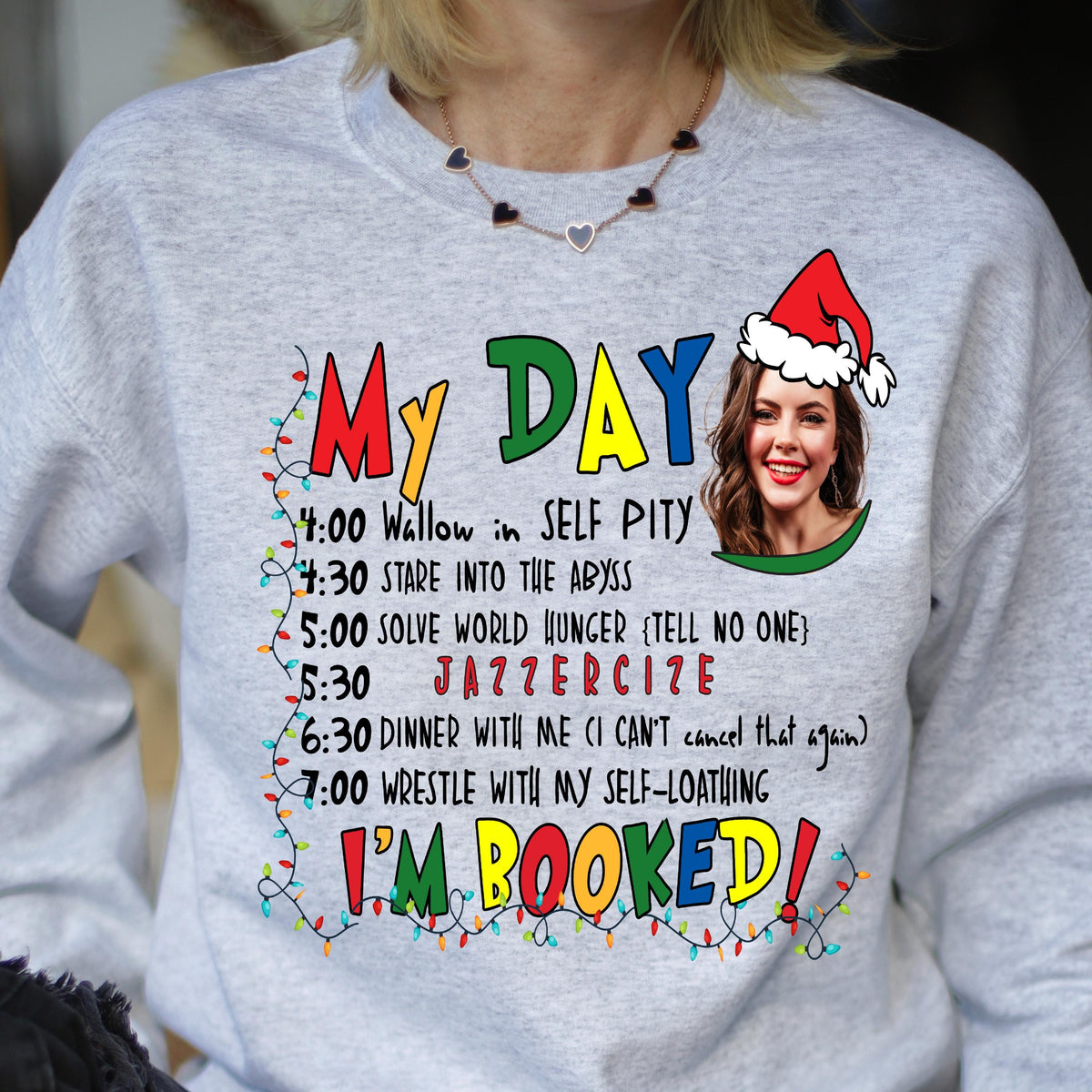 May include: A light gray crewneck sweatshirt featuring a graphic print with the text MY DAY and I'M BOOKED in colorful letters. It includes a schedule of activities, a string of Christmas lights, and a photo of a person wearing a Santa hat.