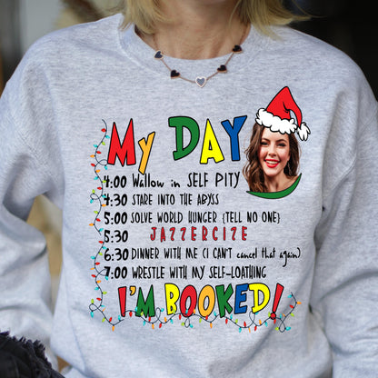 May include: A light gray crewneck sweatshirt featuring a graphic print with the text MY DAY and I'M BOOKED in colorful letters. It includes a schedule of activities, a string of Christmas lights, and a photo of a person wearing a Santa hat.