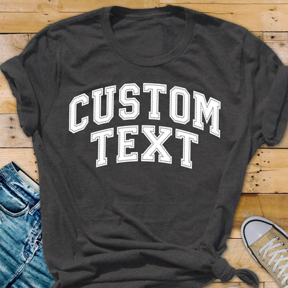 May include: Dark gray t-shirt with the words "CUSTOM TEXT" in white, collegiate-style lettering. The shirt is slightly wrinkled and knotted at the bottom. A pair of blue jeans and a canvas shoe are partially visible.