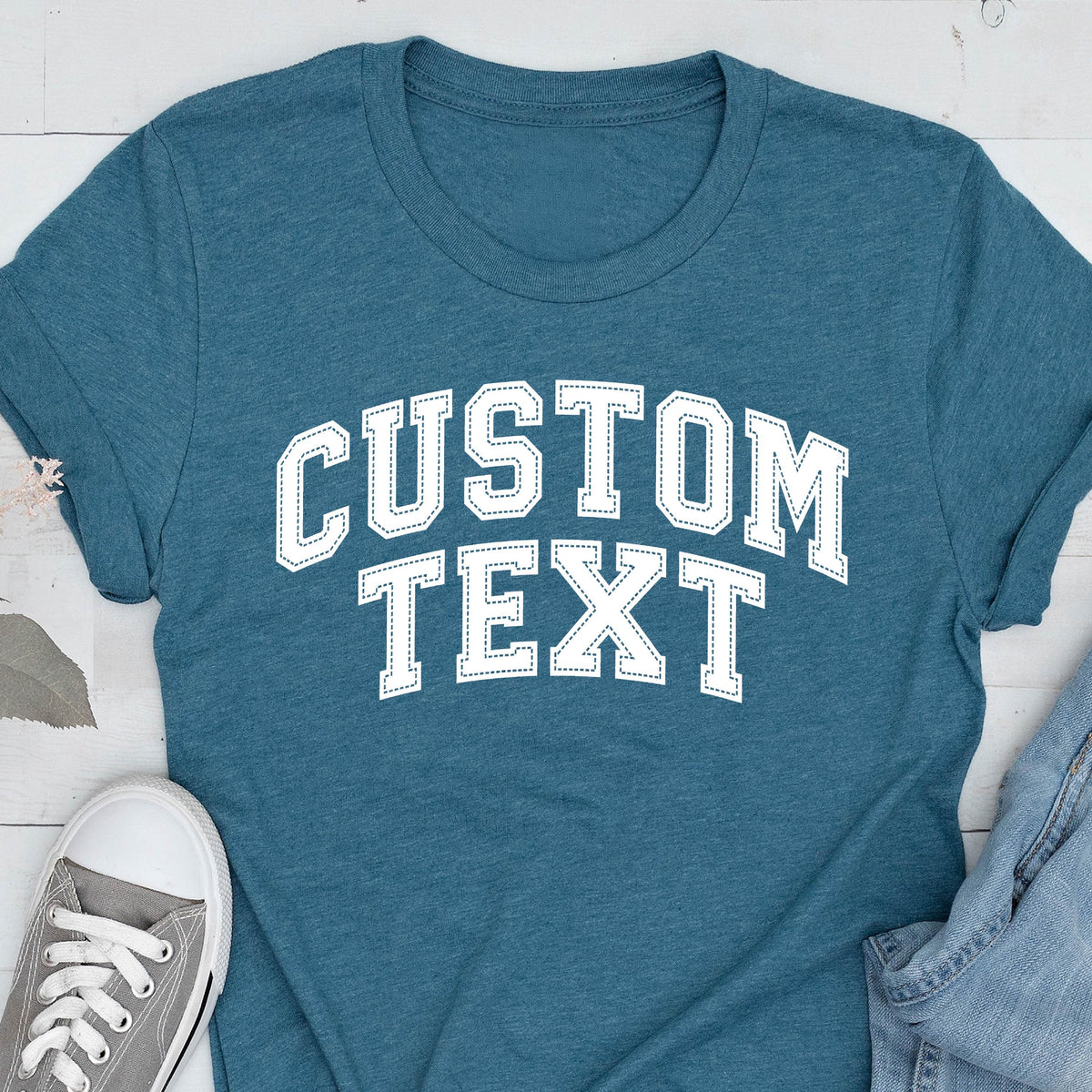May include: Teal t-shirt with the text "CUSTOM TEXT" in white, collegiate-style lettering. The shirt has a crew neck and short sleeves. A gray canvas sneaker and denim jeans are partially visible in the image.