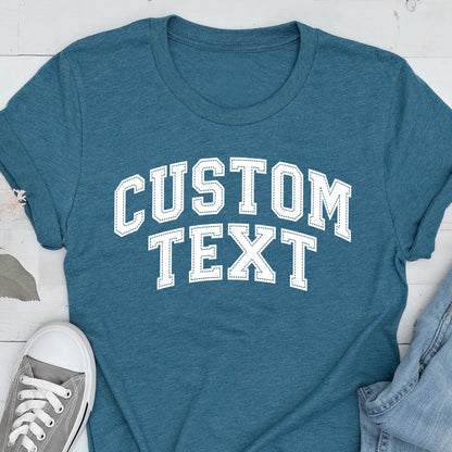 May include: Teal t-shirt with the text "CUSTOM TEXT" in white, collegiate-style lettering. The shirt has a crew neck and short sleeves. A gray canvas sneaker and denim jeans are partially visible in the image.