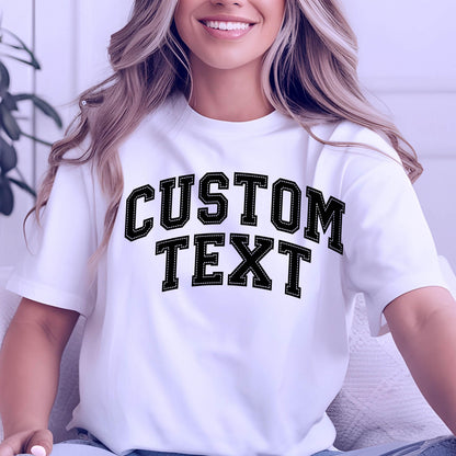 May include: White t-shirt with the text "CUSTOM TEXT" in a black, stitched-style font. The text is in a bold, collegiate-style font. The shirt has short sleeves.